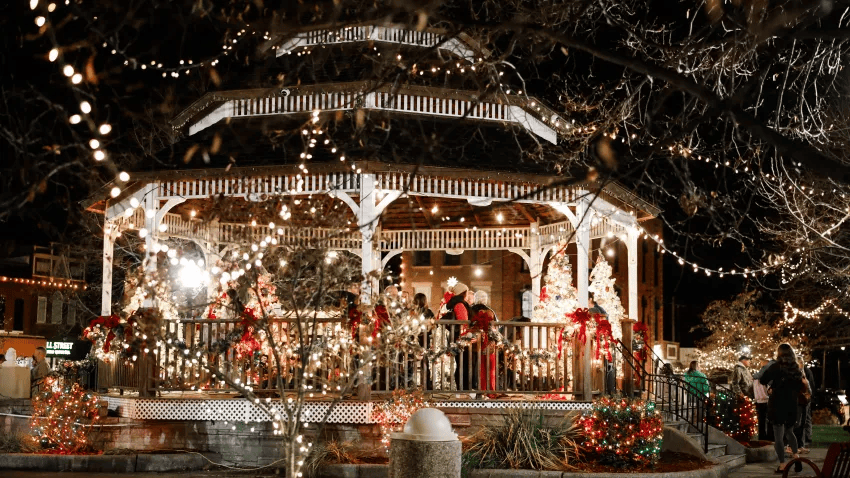 A beautifully decorated gazebo at night, adorned with festive lights and holiday decorations, surrounded by trees and people enjoying the festive atmosphere.