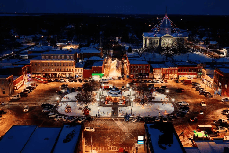 Aerial view of a snow-covered town square illuminated at night, featuring a central gazebo, decorated trees, and surrounding buildings with festive lights.